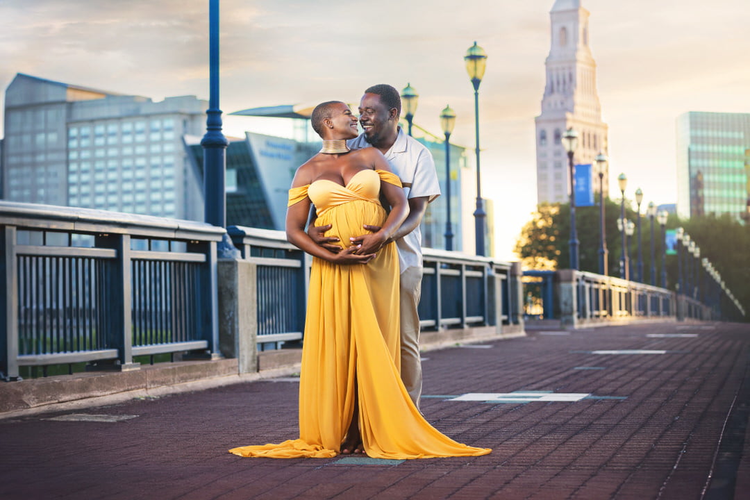 couple with woman in a yellow dress look at each other laughing during their maternity photoshoot in the city
